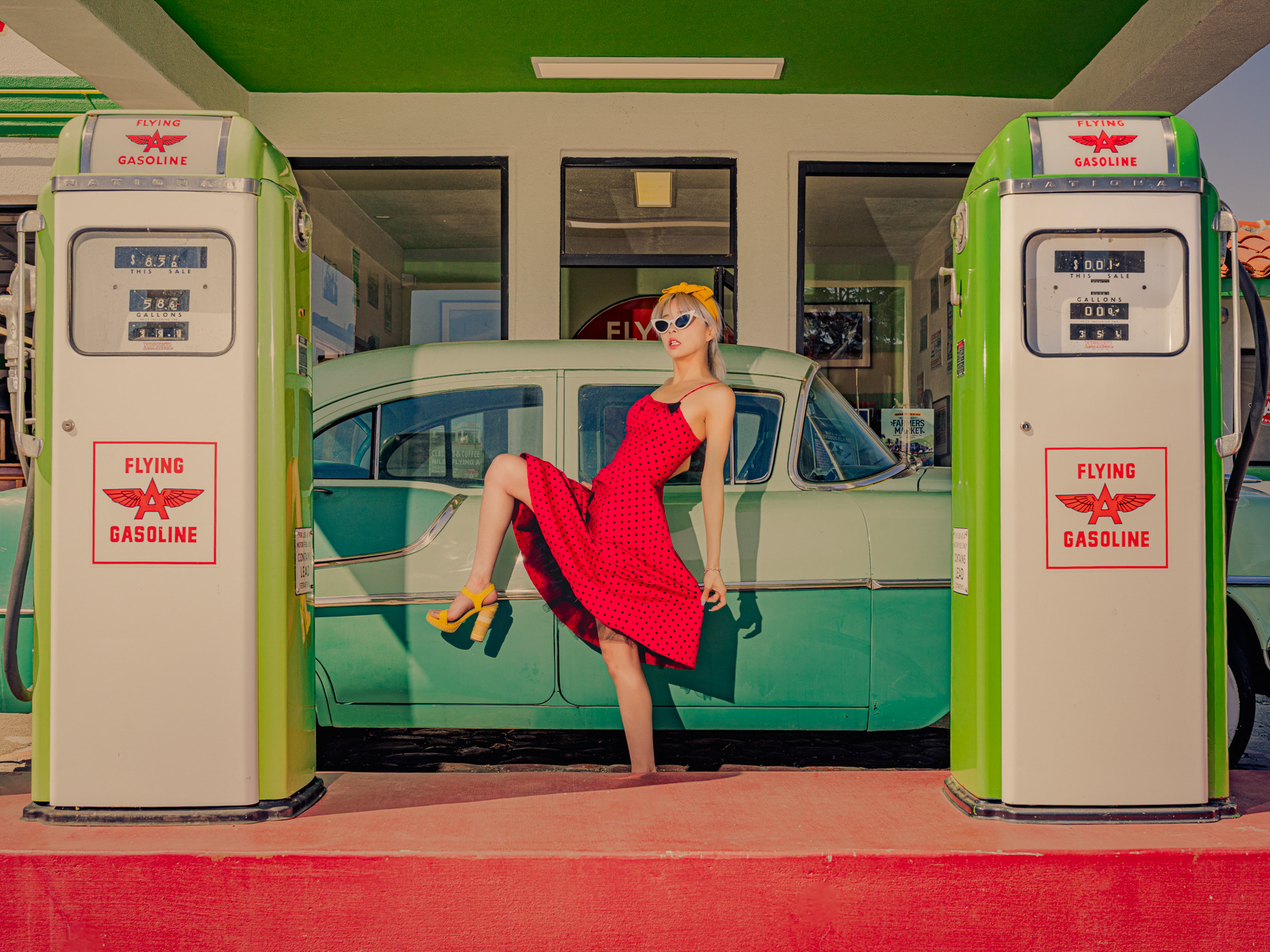 Fashion editorial model in a red dress posed between vintage gas pumps and a classic car